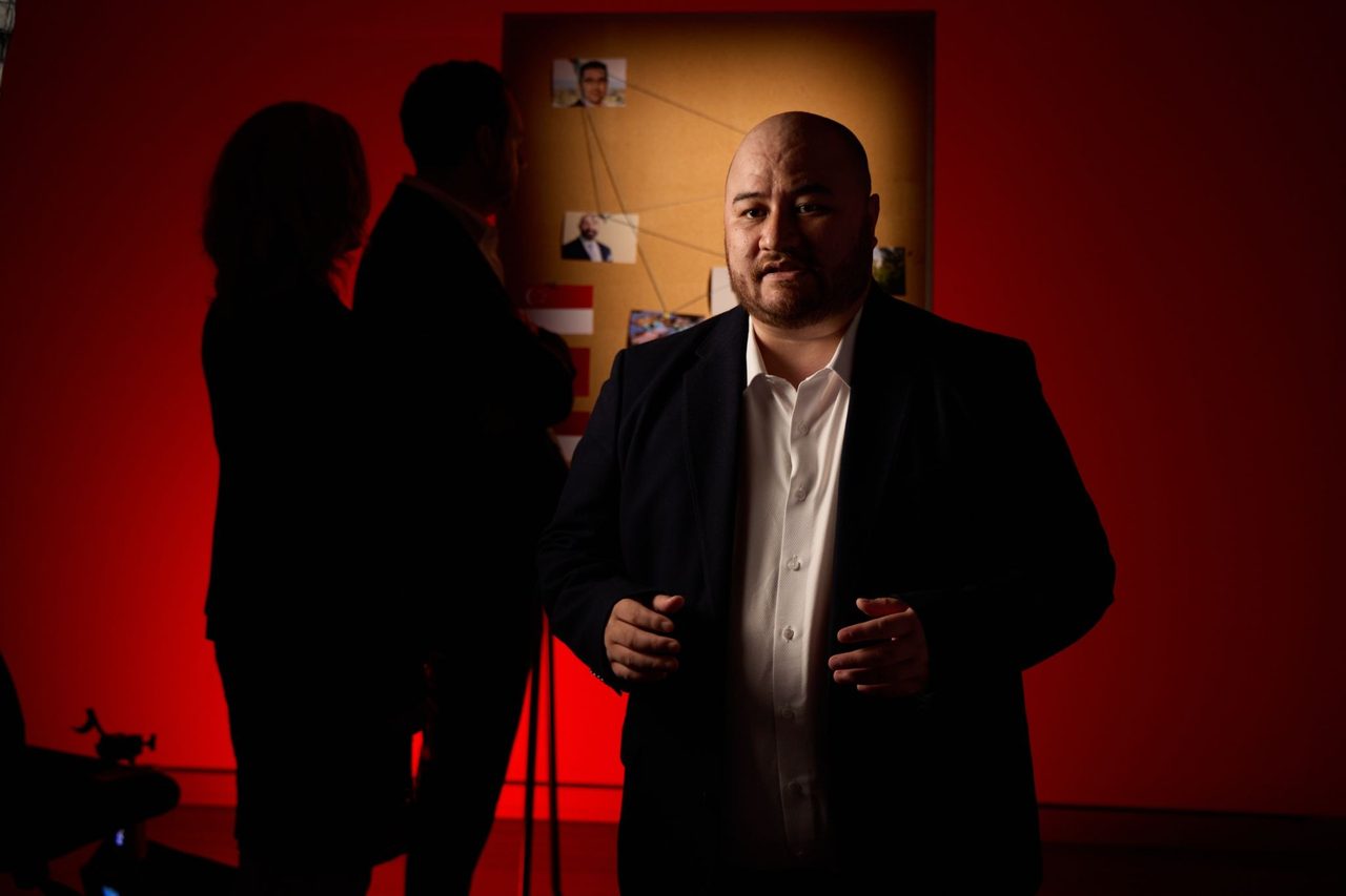 CHOICE Senior Investigative Journalist Jarni Blakkarly tells the story of the collapse of Shield and First Guardian Master Funds while a man and a woman in the background look a cork board with images of people who were involved with the two funds.